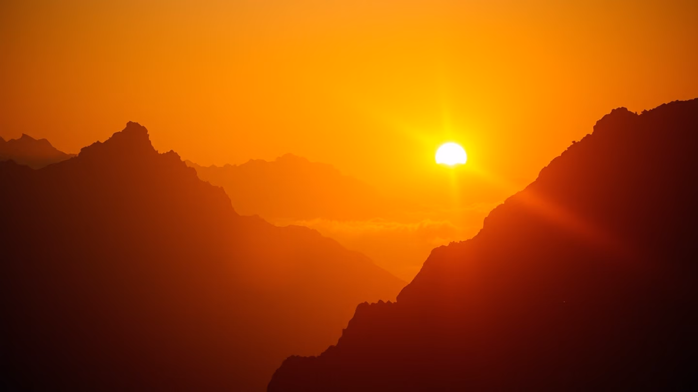 A golden sun rising over the jagged silhouettes of ancient mountain peaks, warm rays piercing through heavy morning fog.