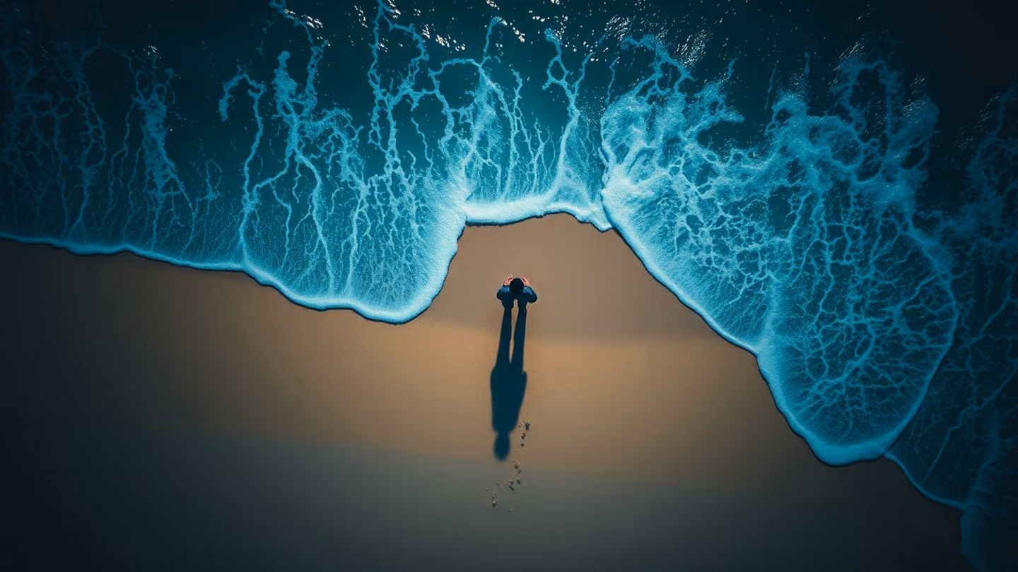Overhead top-down perspective of a lone figure standing exactly where the beige sand meets the deep blue ocean foam, the textures of nature merging, mystical and calm light.