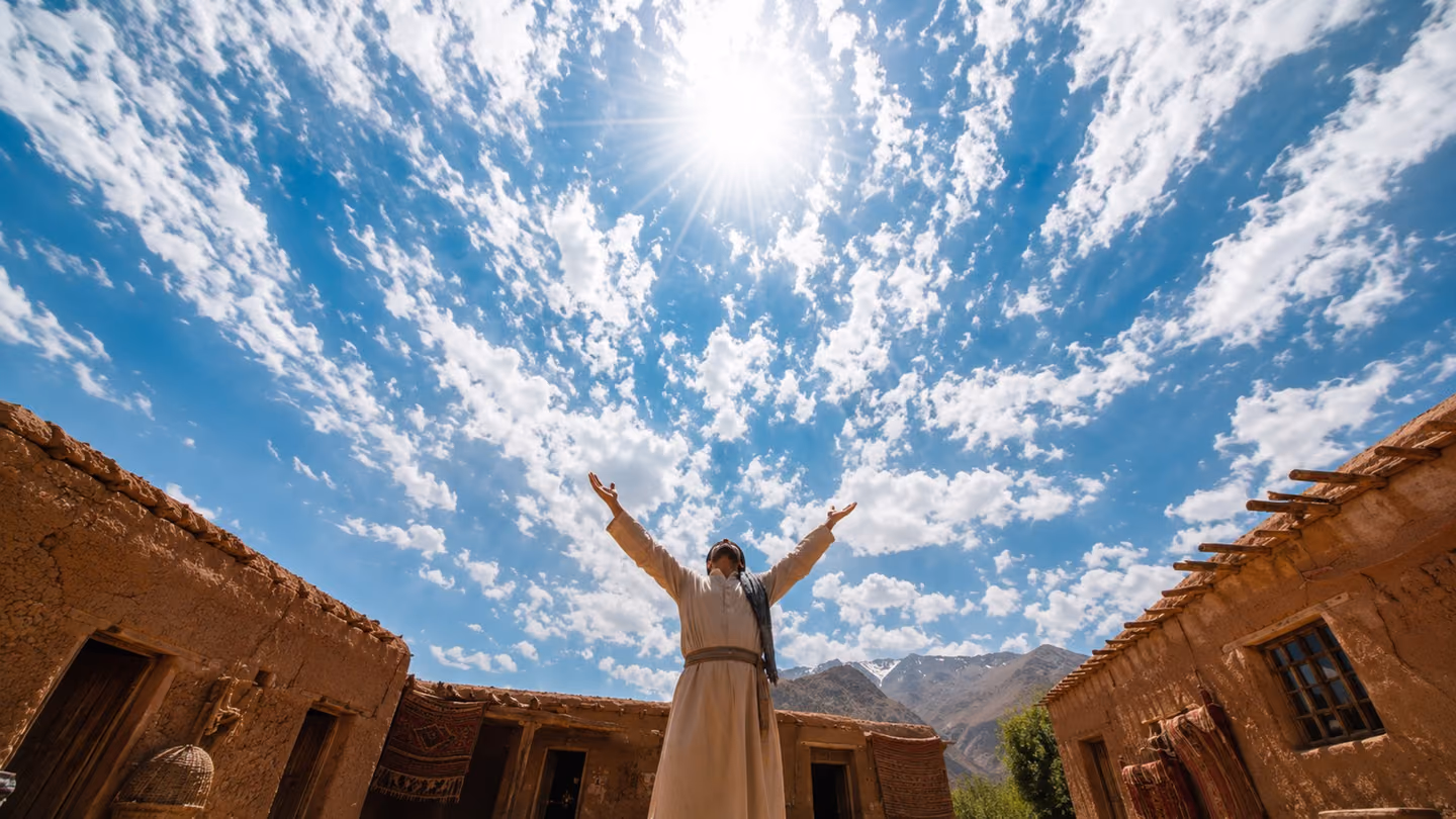 Extreme wide shot looking straight up from a dusty village courtyard in rural Iran: a lone robed Iranian man stands arms flung wide, cracking the vault of a blazing midday sky as rays of light burst outward like shatt…