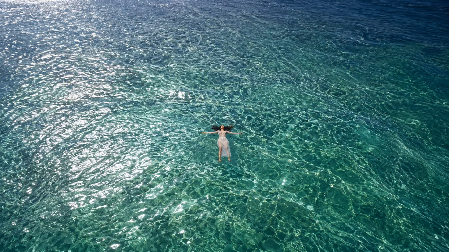 Sweeping overhead wide shot of a vast luminous sea — an Iranian woman floats on her back at the centre, arms open, eyes closed in surrender, her dark hair spreading like ink in the clear water; the ocean surface catch…