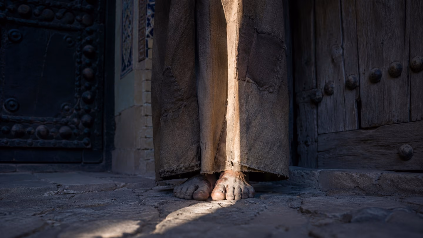 Extreme close-up of bare, dust-covered feet belonging to our solitary Iranian dervish-wanderer — the motif figure — standing perfectly still on ancient cobblestones exactly at the midpoint between two closed doors, on…