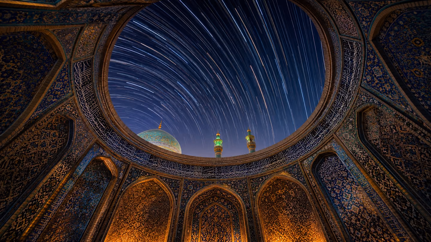 Dramatic slow-tilt wide shot framed through the oculus of a grand Persian dome — looking upward through the circular opening into the night sky, the dome's interior tiled in deep lapis and turquoise geometric patterns…