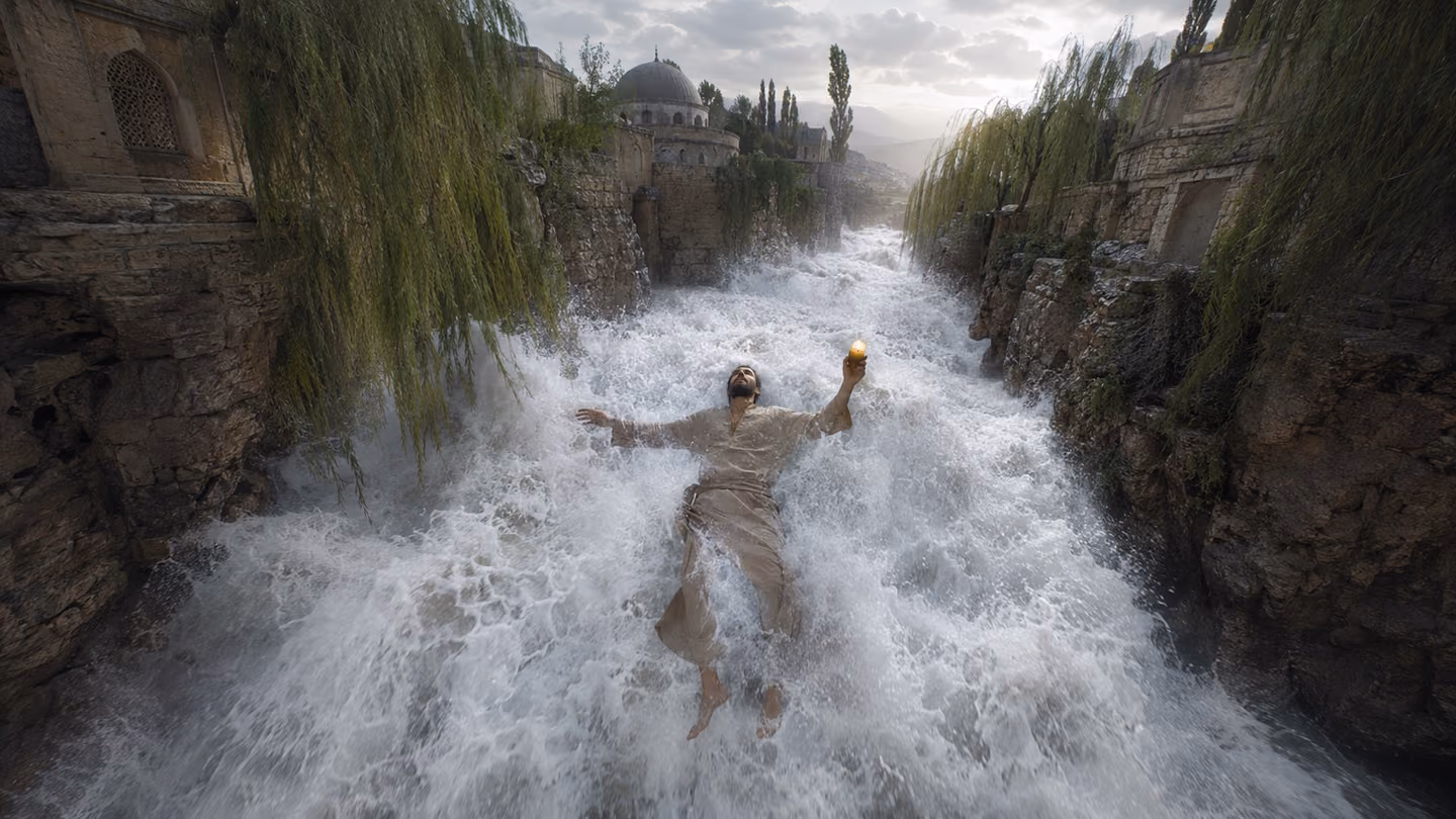 Sweeping wide shot from above — a white flash-flood of foaming water tears through a narrow rocky Persian gorge lined with ancient stone, willows bending to the torrent; the same lean dervish (undyed wool khirqa now s…