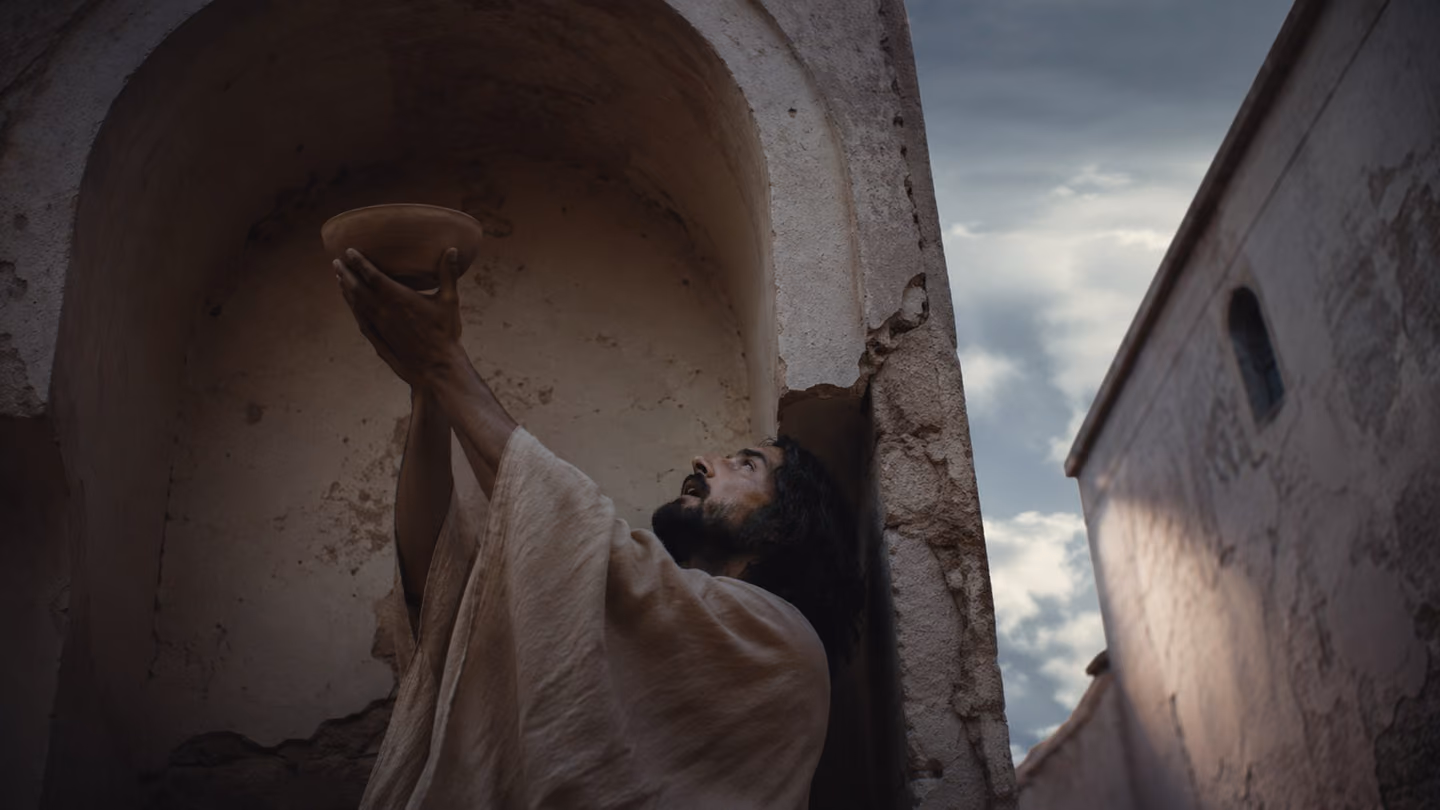 A solitary figure stands in a deep arched alcove cut into the lane wall — shot from below at a steep angle, his hands raising a plain unglazed ceramic bowl toward the arch above him, face tilted skyward in a gesture o…