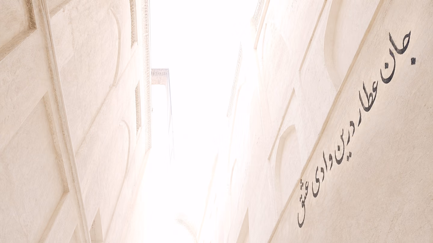 Full white morning — the camera at the floor of the lane looks vertically upward along the tall plaster walls to a sky now blazing with pure white dawn light, and on the right-hand wall a line of Persian calligraphy i…