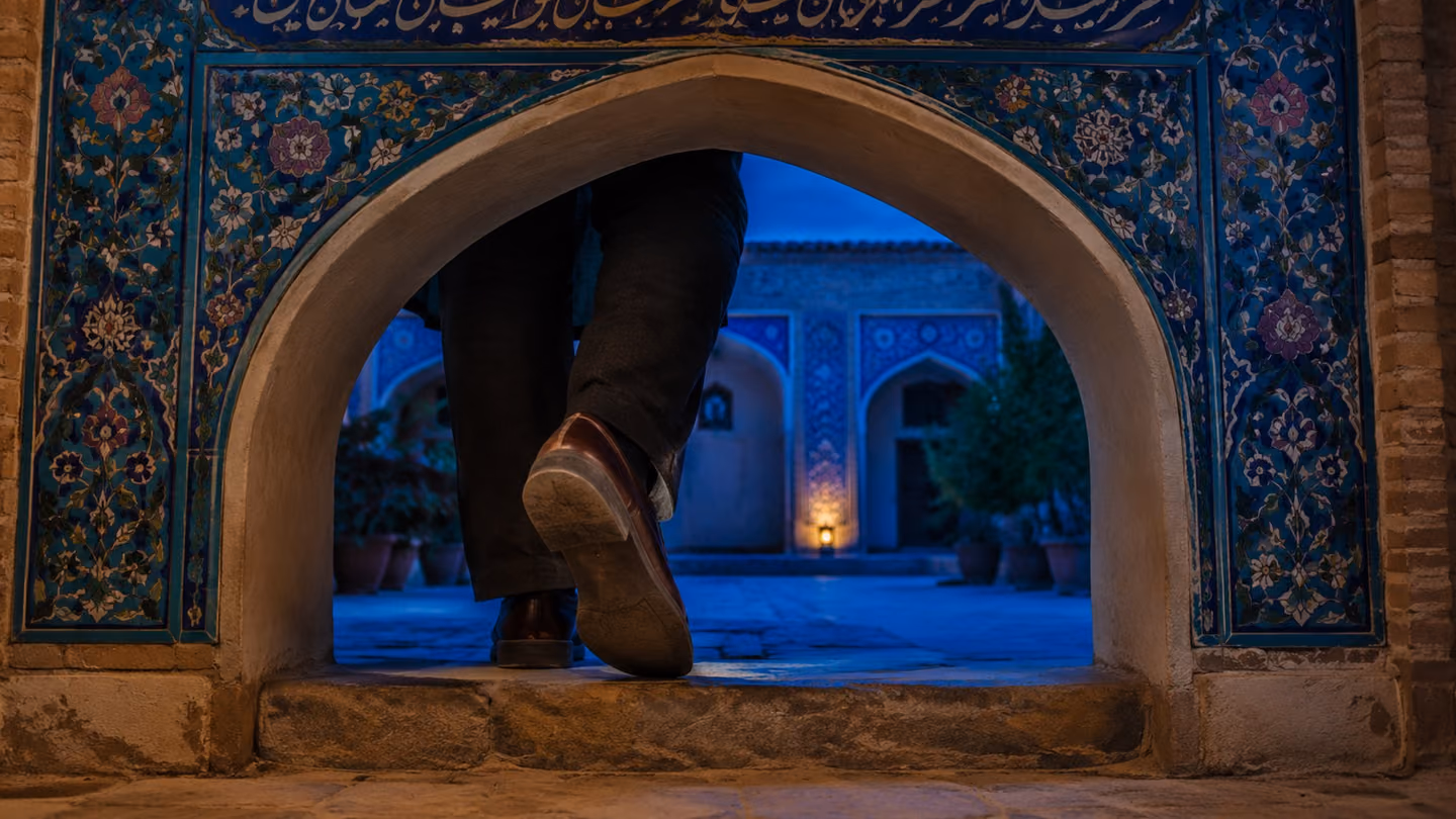 Framed through a turquoise-tiled archway at blue dusk, a man's foot in a plain leather shoe comes down on the threshold of a Kashan courtyard house, mid-stride, entering.