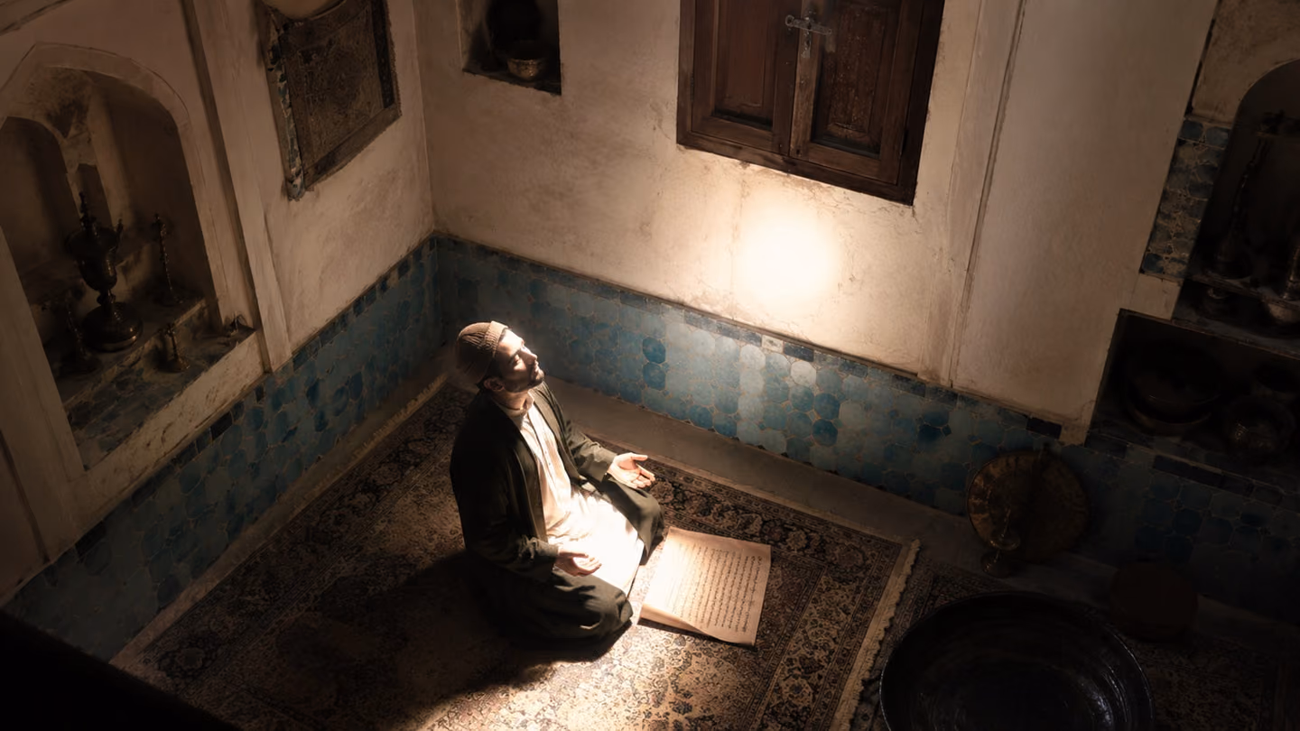 Clear white radiance fills a small Kashan room whose only roof-window is latched shut, the light plainly not coming from it; the householder sits beneath, hands open on his knees, his face lit from in front of him by…