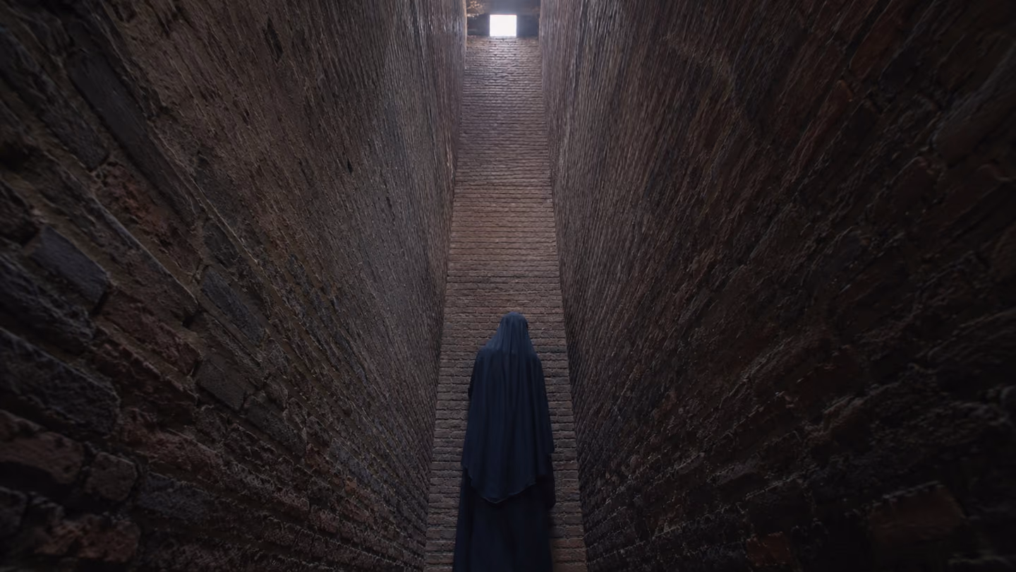 Pale sky shows as a small bright square far up a narrow brick airshaft; at the bottom the young woman stands with her face turned to the bare brick, nothing before her to look at, her hands still.