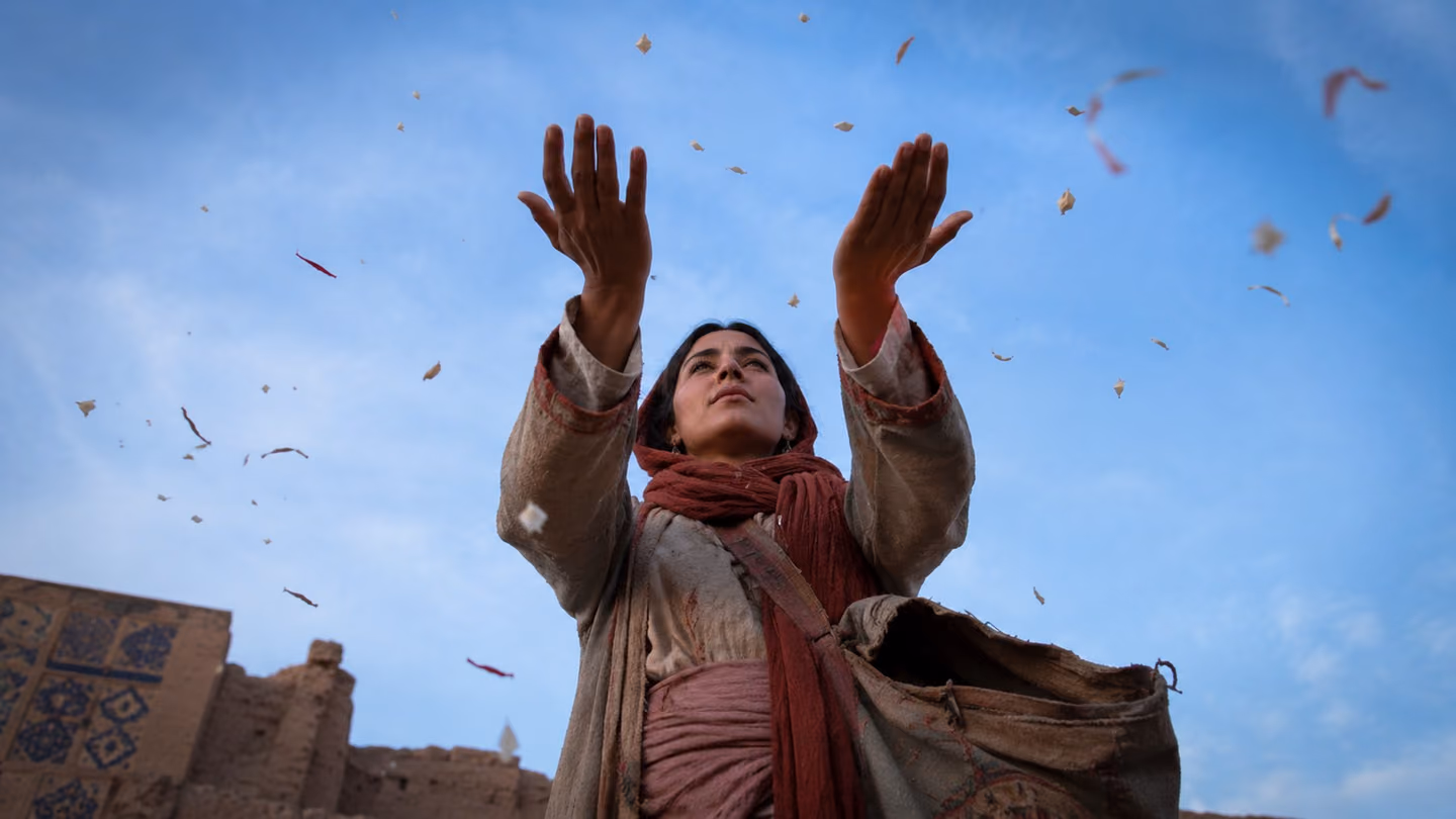 Low from the dust of the road looking steeply up, the woman opens both palms wide above the camera — empty, cupping nothing but the pale blue Khorasan morning.