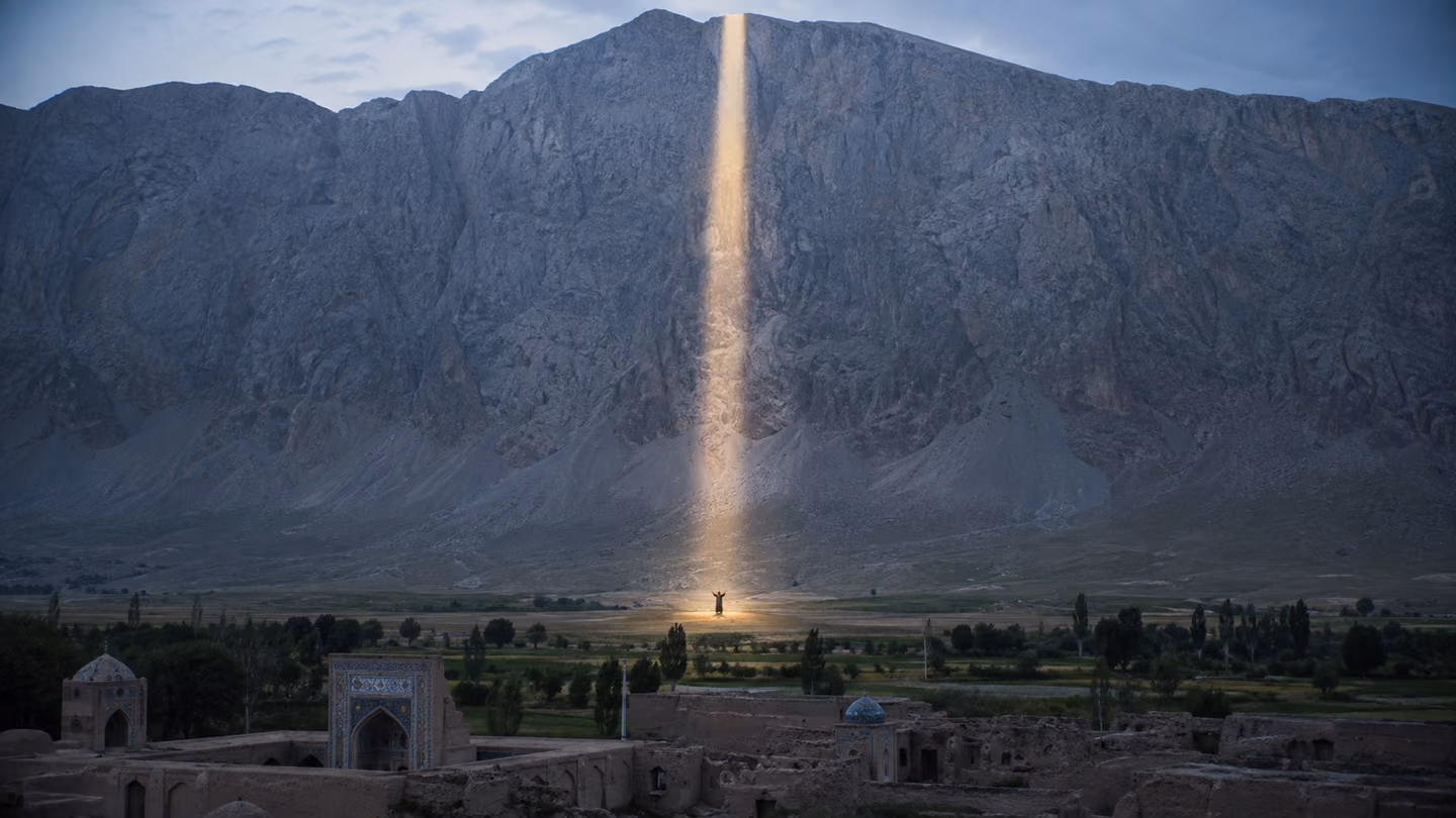 A single clean vertical shaft of risen daylight splits a grey mountain wall above the Khorasan plain from summit to scree; at its foot the woman is very small, arms open and hands empty, holding nothing up between her…