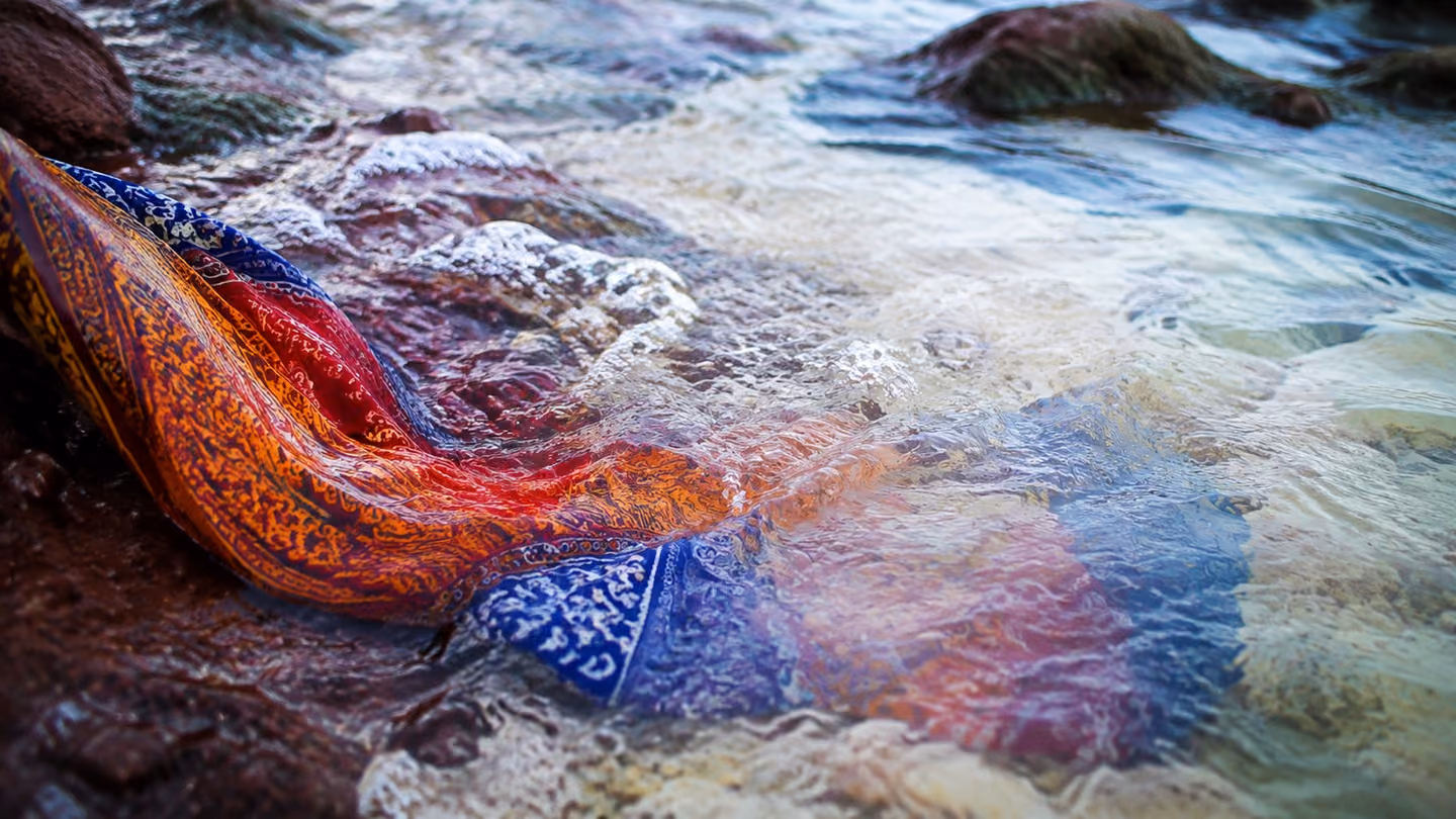 Bandari printed cloth in saffron, crimson and indigo slipping into clear running water, its dye blooming outward in soft ribbons and thinning downstream until the fabric goes pale and translucent.